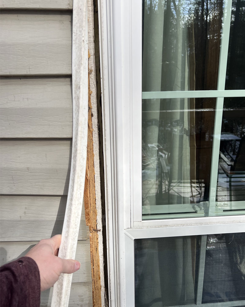 Rotten wood exposed beneath pulled-back siding trim next to a window.