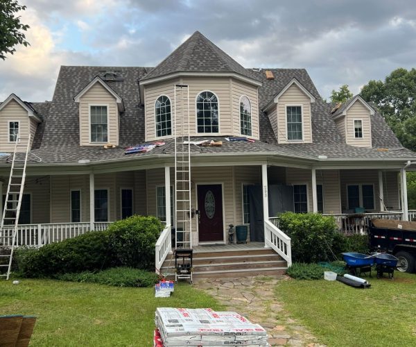 residential-home-roof-replacement-gaf-contractor-columbia-sc Two-story house with tan siding undergoing roof replacement, featuring extension ladders and materials loaded near dormer windows.